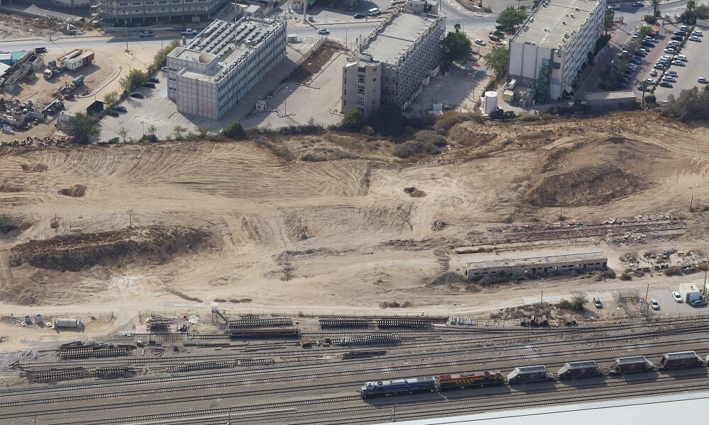 רכבת נוסעת על המסילה, מסביבה חול ובהמשך בניינים